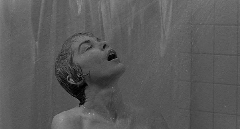 "Black-and-white photo of a person with wet hair standing under a shower, with water streaming down, captured in a tiled bathroom setting."